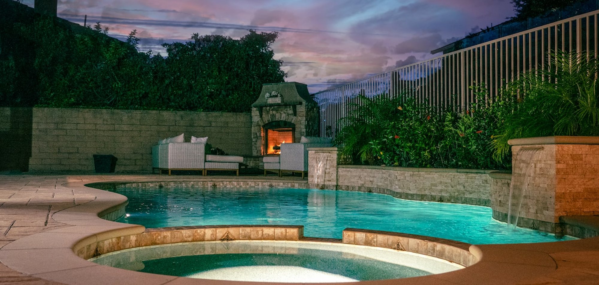 A landscaped pool and patio lit at dusk beside an outdoor fireplace